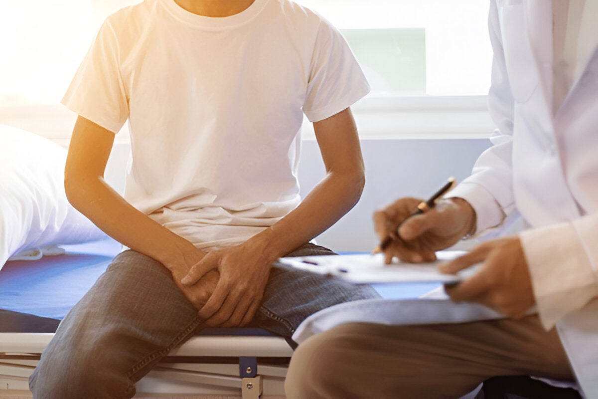 my balls are full the image shows a patient and a doctor. The patient is sitting on the bed with both of his hands covering his testicle area above his pants and the doctor is taking notes