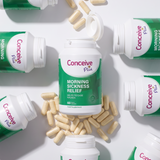 Conceive Plus Morning Sickness Relief dietary supplement bottles and loose beige capsules arranged in a flat-lay composition on a white background.