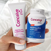 A hand holds a white tube of Conceive Plus Fertility Lubricant and a white bottle of Conceive Plus Men's Fertility Support against a soft white fabric background.