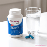 Conceive Plus fertility support supplement bottle with a glass of water on a white surface.