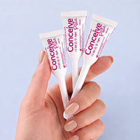 A hand with light pink manicured nails holds three white concealer tubes with pink and purple branding against a soft light blue background. The tubes are labeled 'Conceine Plus' and '12m'.