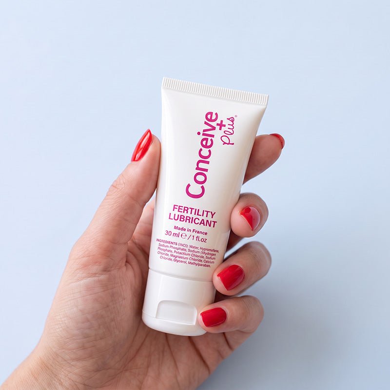 A hand with red-painted nails holds a white squeeze tube of Conceive Plus Fertility Lubricant against a light blue background.