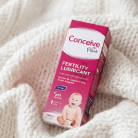 A box of Conceive Plus Fertility Lubricant resting on a soft, white chunky knit blanket. The packaging is predominantly pink and white with text indicating it supports sperm quality and is suitable for pregnant women.